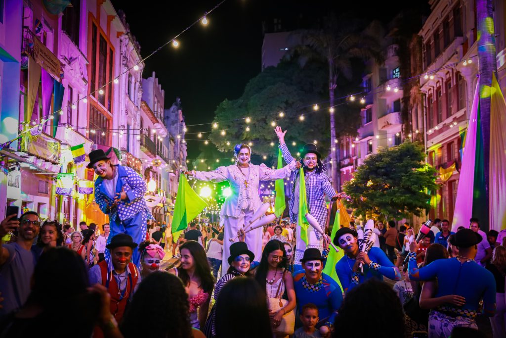 Artistas com fantasias coloridas de Mardi Gras desfilam à noite na Rua da Moeda, no Bairro do Recife, com o público em volta e casario histórico iluminado ao fundo